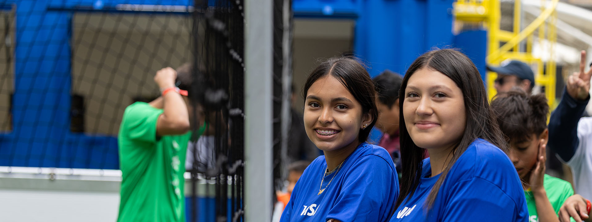 Two young soccer players wearing blue shirts stand next to the street soccer field, looking at the camera.